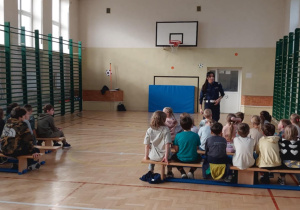 Uczniowie klas I-III na spotkaniu z panią policjantką w sali gimnastycznej. Dzieci z zaciekawieniem słuchają o bezpieczeństwie w ruchu drogowym