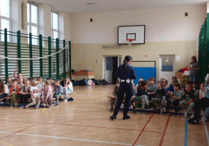 Uczniowie klas I-III na spotkaniu z panią policjantką w sali gimnastycznej. Dzieci z zaciekawieniem słuchają o bezpieczeństwie w ruchu drogowym