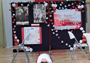 Rozpoczęcie przedstawienia z okazji Święta Niepodległości. Uczennica leży na tle dekoracji z okazji Święta Niepodległości