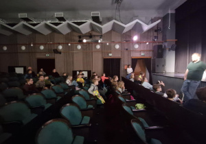 Uczestnicy półkolonii zebrani w sali teatralnej siedzą na krzesełkach i słuchają co mówi pan prowadzący zajęcia