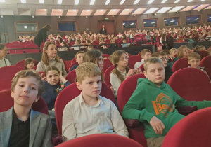 Uczniowie klasy IV wraz z nauczycielem muzyki siedzą w sali teatralnej na fotelach i oczekują na przedstawienie