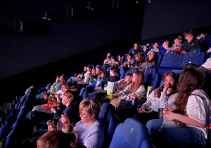 Uczniowie i nauczyciele w sali kinowej na swoich miejscach siedzą i oglądają film pt. ,, Zmiennokształtni 2". Uczniowie oglądają projekcje, chrupiąc popcorn bądź inne przysmaki