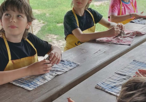 Uczniowie siedzą przy stole pod wiatą, Przed nimi leżą ściereczki a uczniowie ściskają w rękach glinę aby ją uformować.