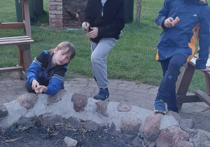 Uczniowie podczas ogniska. Dwóch stoi przy ognisku i patrzy w prawą stronę, jeden kuca przy ognisku dorzucając patyk