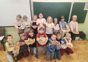 Uczniowie klasy II pozują na tle tablicy, prezentując swoje sałatki, które wykonali na lekcji ,,pod okiem" wychowawcy. Część uczniów stoi a część kuca, wszyscy uśmiechnięci.