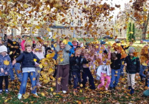 Uczniowie klasy I i III w jesiennym ogrodzie szkoły bawią się liśćmi podrzucając do góry. Wszyscy uśmiechnięci i zadowoleni cudownego dnia spędzonego na świeżym powietrzu