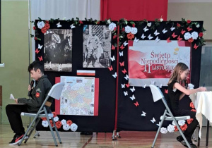 Uczeń i uczennica podczas przedstawienia. Siedzą na krzesełkach plecami do siebie i przemaiwają przez mikrofony