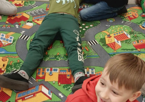 Uczniowie znajdują się na kolorowych matach, leżą na plecach i odgrywają rolę poszkodowanych a inne klęczą obok nich i ćwiczą udzielanie pomocy. Atmosfera jest spokojna i edukacyjna, całe zajęcia mają formę nauki poprzez zabawę, dostosowanej do wieku dzieci.