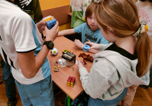 Grupa uczniów stoi wokół ławki, na której rozłożone są różnorodne rebusy i przedmioty do ćwiczenia logiki. Widoczne są kostki Rubika o różnych kształtach, drewniane piramidy oraz metalowe puzzle. Dzieci manipulują przedmiotami, wspólnie szukając rozwiązań.
