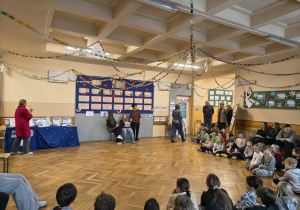 Na korytarzu drugiego piętra zostali zgromadzeni dyrektor, nauczyciele oraz uczniowie naszej szkoły. Osoby dorosłe stoją i obserwują wydarzenie natomiast uczniowie siedzą na podłodze i obserwują prowadzących