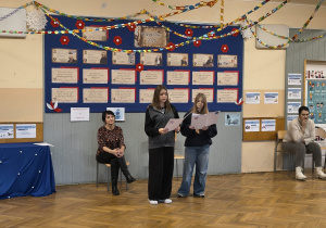 Dwie dziewczyny stoją na środku korytarza i czytają przygotowany tekst. Obok siedzą dorosłe osoby i obserwują występ.