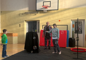 Przedstawienie ,,Zaklinacza baniek". Na zdjęciu Pan Bańka, akcesoria do robienia różnych baniek oraz dwoje uczniów. Chłopiec stoi z boku, natomiast dziewczynka w ręku ma przyrząd do robienia baniek i robi te bańki