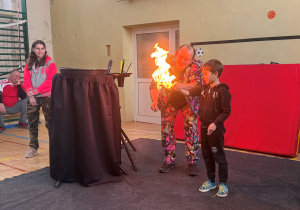 Na zdjęciu Pan Bańka, a przy nim chłopiec, który ma wystawioną rękę do przodu i z jego ręki zdobywa się ogień. Po lewo stoi dziewczynka i przygląda się przedstawieniu