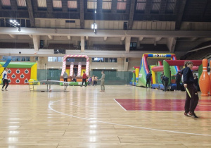 Dzieci na hali sportowej MOSiR, dzieci są rozproszone po różnych stanowiskach, są przy dwóch dmuchańcach, dwoje dzieci gra w ping-ponga, chłopiec stoi przy ogromnym kręglu