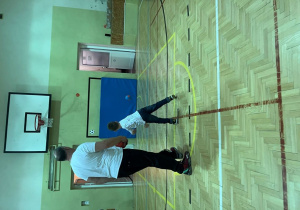 Sala gimnastyczna, uczestnik wraz z wychowawcą odwróceni tyłem do aparatu. Pan kontroluje czy uczestnik prawidłowo rzuca do celu.