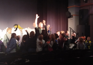 Przy scenie teatru, stoją wszyscy uczestnicy półkolonii a przed nimi prowadzący warszaty wybiera chętnych do przeprowadzenia csenki