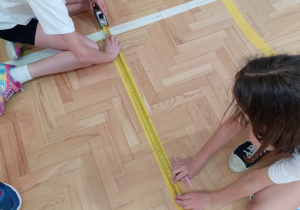 Praca w terenie z długą taśmą mierniczą – uczniowie wspólnie mierzą dystanse na parkiecie, przygotowując dane do obliczeń.