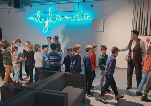 Uczniowie w Teatrze Pinokio ustawieni w parach pod neonowym napisem Nibylandia