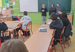 Uczniowie klasy ósmej siedzą w ławkach i wysłuchują ofert edukacyjnej jednej ze szkół ponadpodstawowych. Pani przy tablicy przedstawia ofertę szkoły którą reprezentuje
