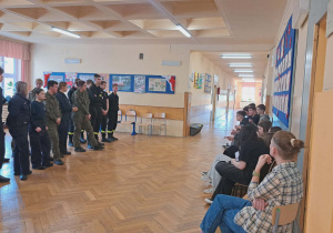 Na korytarzu drugiego piętra klasa VIII spotkała się z uczniami szkoły ponadpodstawowej ,,Oświatowiec". Uczniowie wspomnianej szkoły ubrani w wojskowe mundury stoją na baczność przed uczniami klasy VIII, którzy siedzą na krzesłach