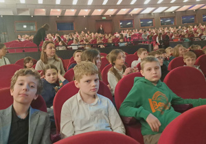 Uczniowie klasy IV wraz z nauczycielem muzyki siedzą w sali teatralnej na fotelach i oczekują na przedstawienie