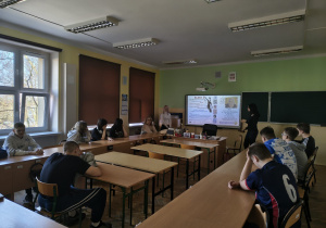 Uczniowie klasy VIII siedzą w ławkach w sali. Na tablicy multimedialnej wyświetlona jest prezentacja i uczennice z XXXIV Liceum Ogólnokształcącego przedstawiają ofertę