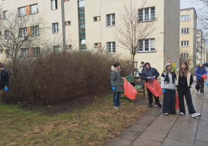 Uczniowie naszej szkoły sprzątają teren niedaleko szkoły. Mają czerwone worki, do których wrzucają znalezione śmieci obok roślinności i bloku przy ulicy Perla.