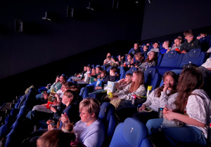 Uczniowie i nauczyciele w sali kinowej na swoich miejscach siedzą i oglądają film pt. ,, Zmiennokształtni 2". Uczniowie oglądają projekcje, chrupiąc popcorn bądź inne przysmaki