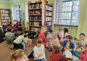 Uczniowie klas I i II siedzą na wykładzinie w bibliotece i jedzą śniadanie