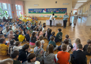 Zgromadzeniu uczniowie na korytarzu szkoły w tle napis Wesołego Alleluja oraz prace uczniów. Przy stołach stoją organizatorzy konkursów i nagrody.