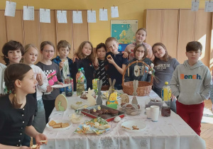 Uczniowie klasy 3 zgromadzeni przy swoim wielkanocnym stole, pozując do zdjęcia z uśmiechami na twarzy. W tle wiszą karty pracy wykonane przez ucznió