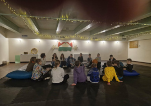 Teatr Arlekin - sala ćwiczeń - przytłumione światło. Uczniowie siedzą w kole na pufach i słuchają Pani prowadzącej warszaty.