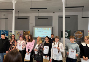 Uczennica klasy VI na wręczeniu nagród za udział w Międzyszkolnym konkursie plastycznym pt. ”Łódź- miasto z nici utkane”. Uczennica pozuje wśród innych finalistów w dużej auli.