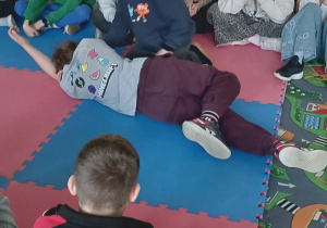 Uczniowie klas 1-3 na zajęciach w świetlicy szkolnej poznają tajniki pierwszej pomocy podczas różnych sytuacji zagrażających życiu innych osób. Na zdjęciu uczniowie siedzą w kręgu a dwóch z nich przedstawia pozycję boczną bezpieczną.