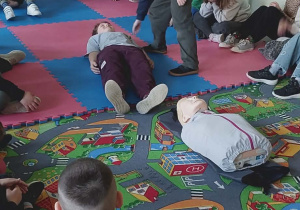 Uczniowie klas 1-3 na zajęciach w świetlicy szkolnej poznają tajniki pierwszej pomocy podczas różnych sytuacji zagrażających życiu innych osób. Na zdjęciu uczniowie siedzą w kręgu dwóch uczniów prezentuje jak sprawdzić czy z leżący człowiek ma kontakt.