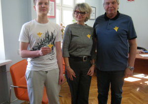 Uczeń klasy VII pozuje na zdjęciu z panią z sekretariatu oraz dyrektorem szkoły w gabinecie u dyrektora. Wszyscy mają przypięte do ubrań papierowe żonkile.