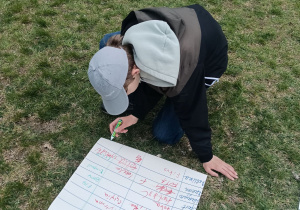 Widok z góry na ucznia klasy 6 w szarej czapce z daszkiem. Chłopak klęczy na trawie i pisze zielonym mazakiem na dużym arkuszu papieru.