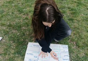 Widok z góry na kucającą dziewczynę. Uczennica jest pochylona nad kartonem z tabelą leżącym na trawie i pisze w nim długopisem.