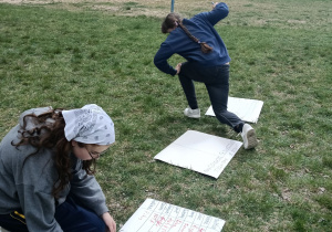 Dwie uczennice na trawniku przed szkołą. Jedna klęczy i pisze mazakiem na dużym kartonie, druga stoi kawałek dalej w pozie startowej. Zajęcia odbywają się na zielonym terenie szkoły.