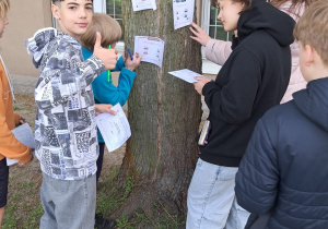 Grupa uczniów z klasy 5 czyta zadanie matematyczne przyczepione do pnia drzewa. Jeden z chłopców uśmiecha się i pokazuje kciuk w górę.