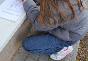 Uczennica kuca na zewnątrz budynku i w skupieniu rozwiązuje zadanie matematyczne, opierając kartkę o parapet okna.