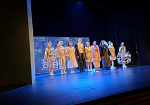 Scena teatru oświetlona niebieskim światłem. Na scenie stoją aktorzy w kostiumach scenicznych użytych podczas przedstawienia, aktorzy żegnają się z publicznością.