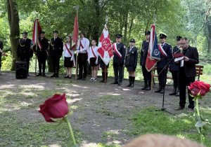 Park im. Józefa Piłsudskiego, uczniowie klasy siódmej stoją ze sztandarem szkoły przy kamieniach upamiętniających wydarzenia sprzed lat. Obok uczniów przemawia wojskowy