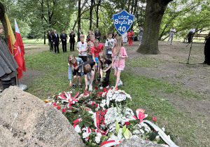Uczniowie klasy czwartej z szyldem szkoły stawiają znicz przed kamieniami upamiętniającymi rewolucjonistów w parku i. J. Piłsudskiego. W tle wojskowi oraz harcerze i osoby reprezentujące miasto Łódź.