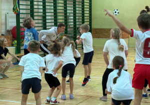 Uczniowie na sali gimnastycznej wraz z zawodnikiem Łódzkiego Klubu Sportowego wykonują przysiady z wyskokiem.