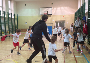 Uczniowie na sali gimnastycznej wraz z zawodnikiem Łódzkiego Klubu Sportowego bawią się w berka.