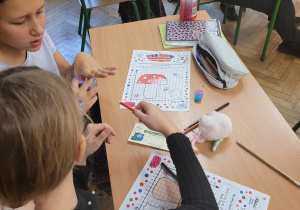 Dwie dziewczynki siedzą w szkolnej ławce i skupione rozwiązują zadanie na karcie pracy zatytułowanej "Dzień Kropki". Jedna z nich trzyma w ręku kredkę, a na ławce leżą przybory szkolne i podręcznik. W tle widoczni są inni uczniowie.