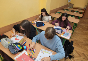 Cztery uczennice siedzą w szkolnych ławkach, pracując z zadaniami matematycznymi w układzie współrzędnych. Na stolikach widoczne są piórniki, kredki i podręczniki.