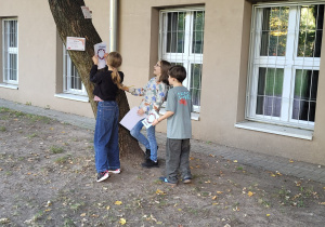 Troje uczniów stoi przy drzewie obok budynku szkoły. Na pniu umieszczone są kartki ze stacjami zadaniowymi. Uczniowie wspólnie odczytują zadanie matematyczne i zapisują odpowiedzi na swoich kartach.