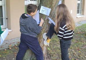 Dwoje uczniów stoi przy drzewie obok budynku szkoły. Na pniu umieszczona jest karta oznaczona jako „Stacja 9” z zadaniem matematycznym. Uczniowie odczytują zadanie i zapisują rozwiązanie na swoich kartach pracy.